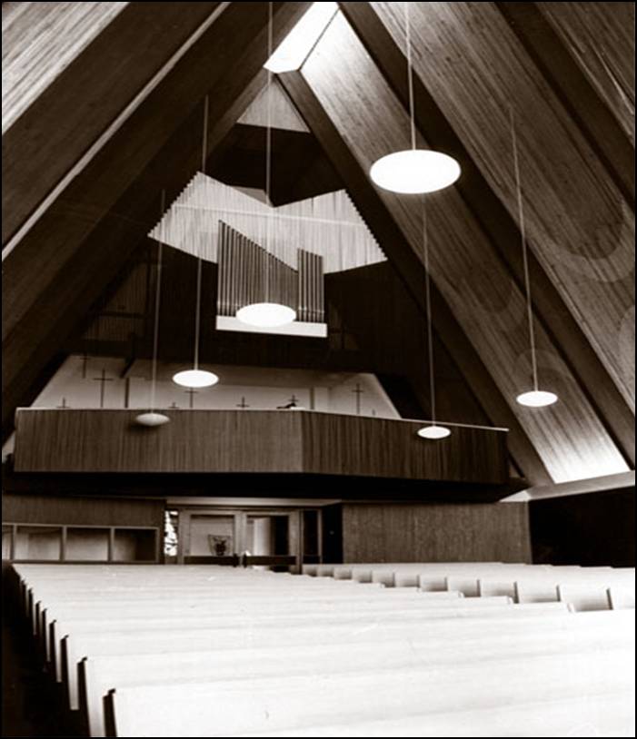1964 historic photograph of the interior sanctuary of Trinity Lutheran Church in Richmond, BC, looking south. The image features a balcony organ loft with a pipe organ, modern pendant lighting, and rows of pews. The steep A-frame ceiling is finished in wooden acoustic battens. The main sanctuary entrance is visible below the organ loft.