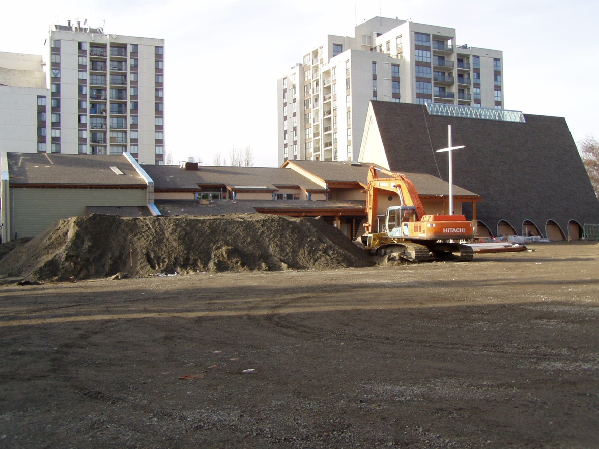 A 2003 photograph showing the completed exterior of the new multi-use facility at Trinity Lutheran Church in Richmond, BC. The image features the new roofline integrated with the 1964 A-frame sanctuary, an orange excavator working on site grading, and a large pile of preloading in the foreground.