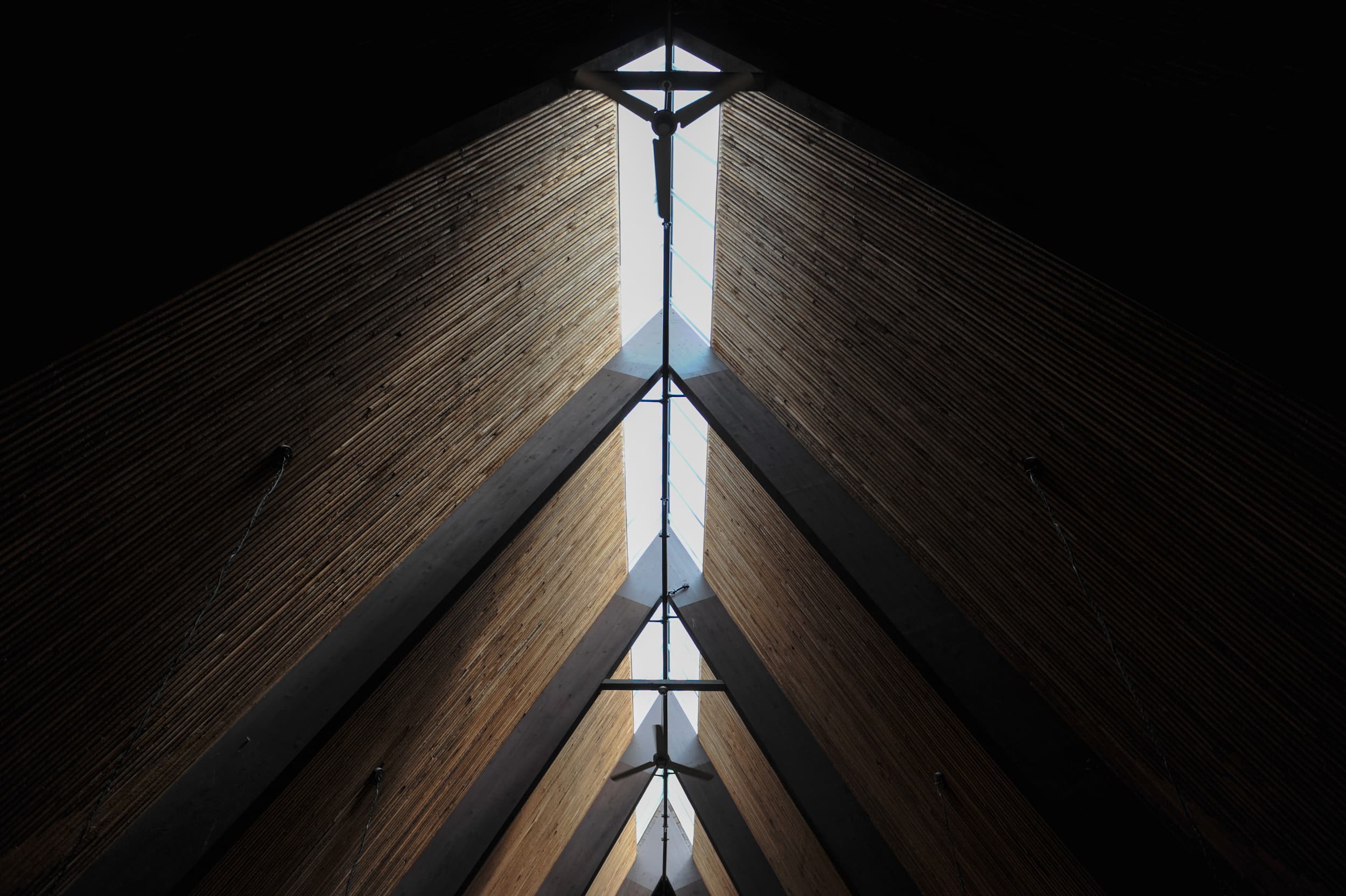 A dramatic architectural perspective looking directly up at Trinity Lutheran Church’s high, vaulted A-frame interior ceiling. A narrow skylight runs the length of the ridge, casting a bright line of natural light that bisects the steeply pitched ceiling. The repeating patterns of dark structural beams and warm wooden slats create a sense of symmetry and soaring vertical space.