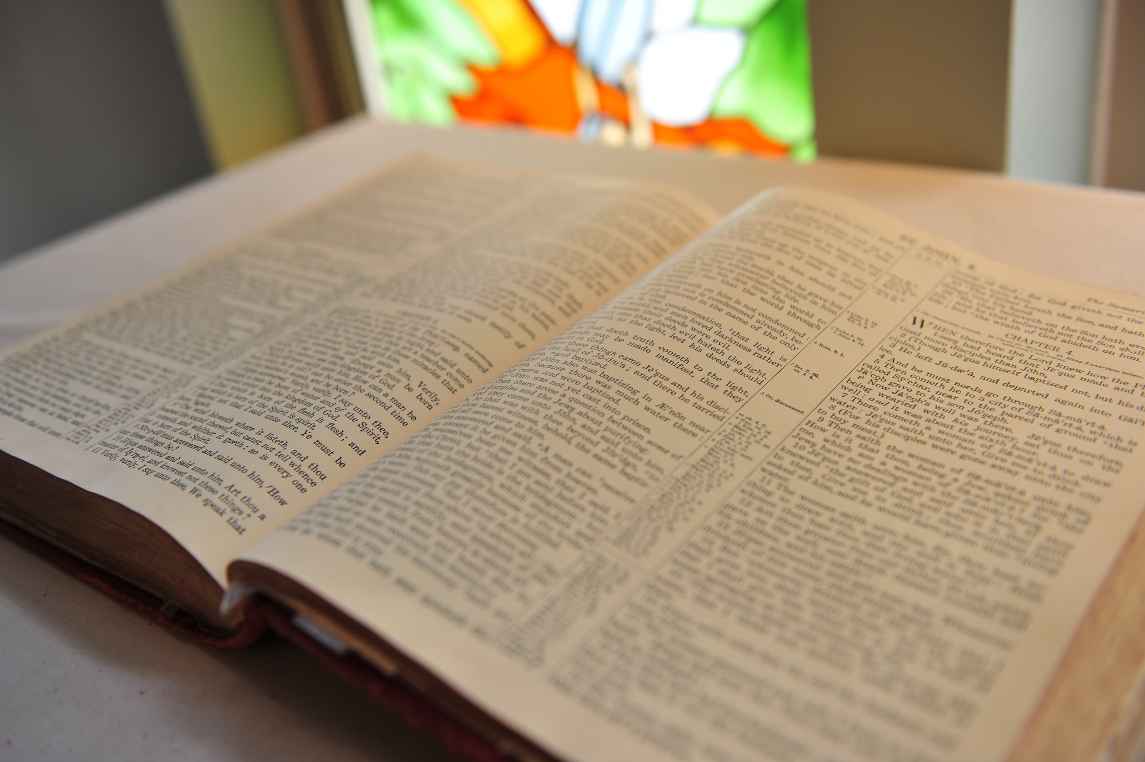 A close-up of an open Bible resting on the altar in the Trinity Lutheran chapel, confirming the congregation's belief in the inerrant Word of God. In the background is a stained-glass window depicting the Trinity and scenes of Law and Gospel.